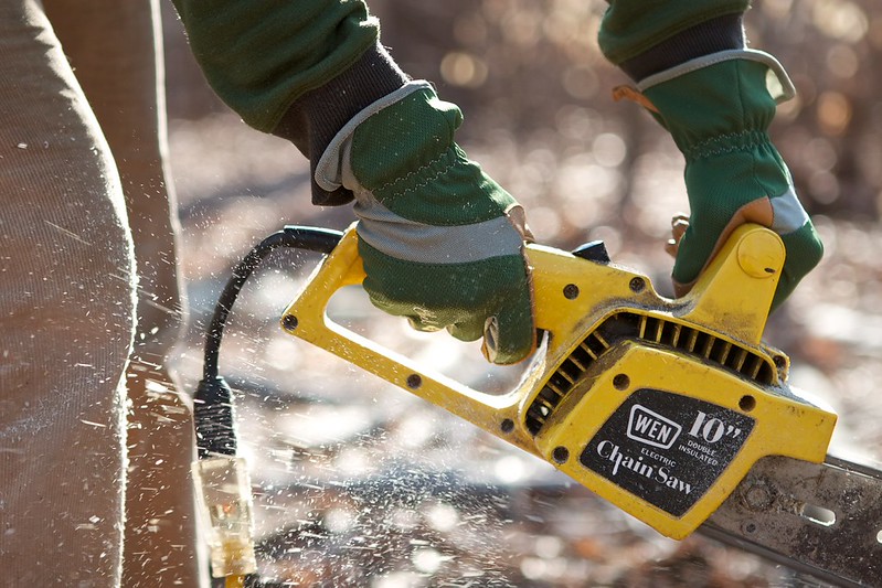 A photograph of hands wearing work gloves using an electric chain saw.