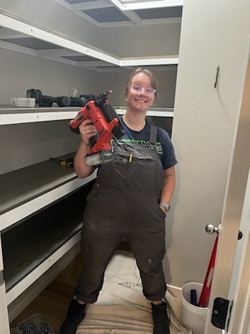Virginia Phelps holding a nail gun and smiling at the camera. Virginia is a white person with glasses.