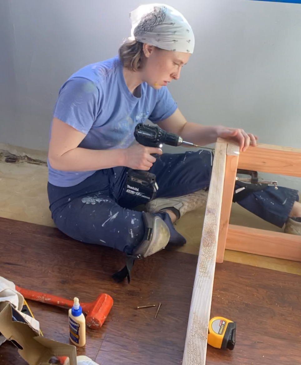 Jess Crask in profile, using a drill to assemble furniture. Jess is a white woman with a white bandana over her hair.