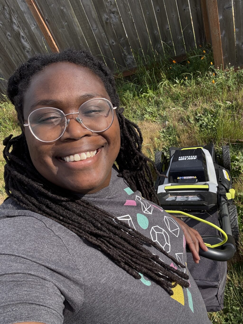 Kera Nellor smiling at the camera with a lawn mower in the background. Kera is a Black woman with long locs and glasses.